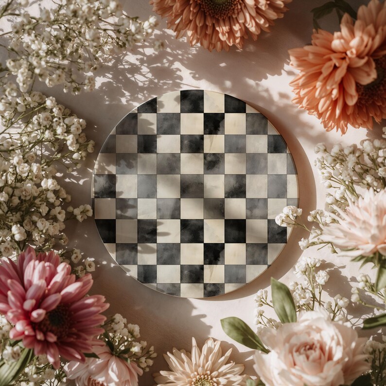 Op de afbeelding: Een ronde decoratieve plaat met een zwart-wit geblokt patroon. De plaat is omgeven door roze en perzik bloemen en witte gipskruid, waardoor een zacht, bloemenframe ontstaat.
