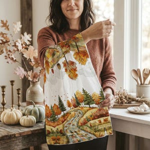 Puede incluir: Un paño de cocina blanco con una ilustración en acuarela de un paisaje otoñal. El diseño presenta un camino sinuoso, árboles y hojas caídas en tonos naranja, amarillo y verde. El paño es sostenido por una persona.