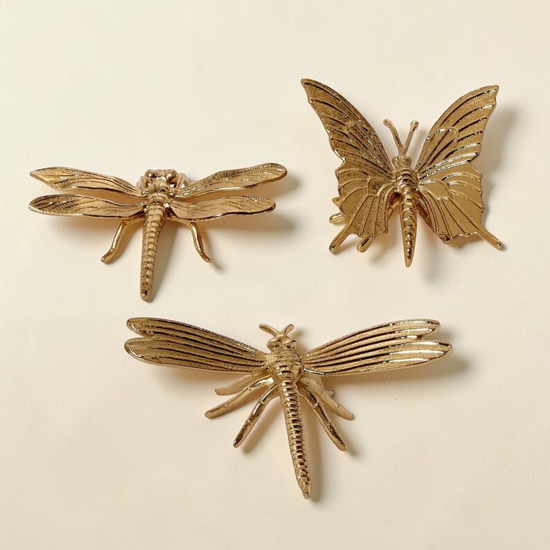May include: Three gold-colored insect and butterfly decorative objects. The insects are dragonflies, and the butterfly has detailed wing patterns. The objects are displayed on a light-colored surface.
