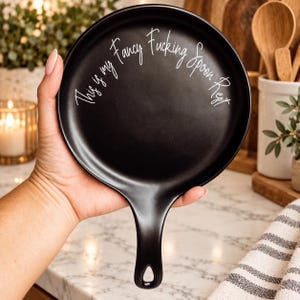 May include: A round, matte black ceramic spoon rest with a handle featuring a hole for hanging. The surface is printed with the white cursive text, "This is my Fancy Fucking Spoon Rest," arranged in a circular arc along the upper edge.
