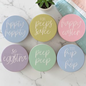 May include: Six pastel-colored round coasters with white cursive text. The coasters are light blue, yellow-green, pink, lavender, mint green, and light blue. Text includes "hippity hoppity", "for peeps sake", "happy easter", "so eggstra", "peep peep", and "too hip to hop".