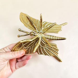 May include: A gold-colored butterfly-shaped decorative object. The butterfly has detailed wing patterns and antenna. The object is held by a hand, showcasing its size and design. The butterfly is made of metal.