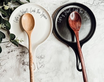 May include: Two ceramic spoon rests, one white and one black, with the text "Life is Short... Lick the Spoon" on each. The white spoon rest has a wooden spoon with the text "Homemade with love, I've licked the spoon" engraved on it. The black spoon rest has a dark wood spoon.