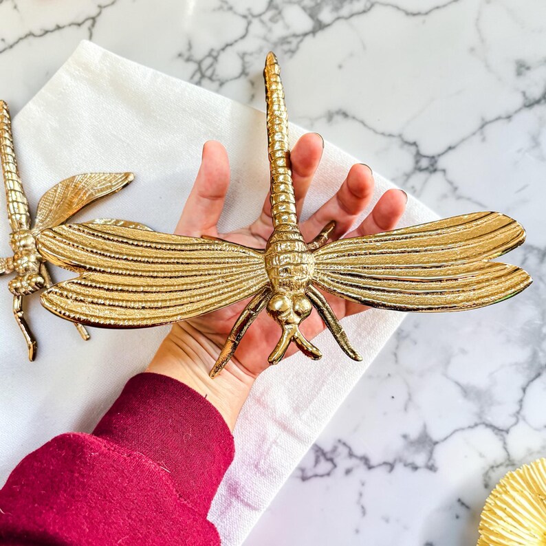 May include: Two gold-colored dragonfly sculptures. The dragonflies have detailed wings and bodies, with a textured finish. The sculptures are displayed on a white cloth, with a marble-patterned surface in the background. The dragonfly sculptures are decorative.
