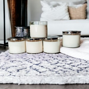 May include: Five white candles in glass jars with silver lids are arranged in a pyramid shape on a patterned rug. The candles are all the same size and have a creamy white wax.