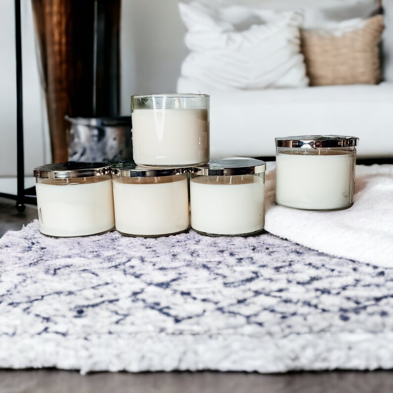 May include: Five white candles in glass jars with silver lids are arranged in a pyramid shape on a patterned rug. The candles are all the same size and have a creamy white wax.