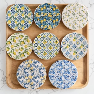 Puede incluir: Nueve posavasos redondos de cerámica con varios diseños de limones y flores. Los posavasos presentan limones amarillos, hojas verdes y patrones florales azules y blancos. Están dispuestos sobre una bandeja de madera.