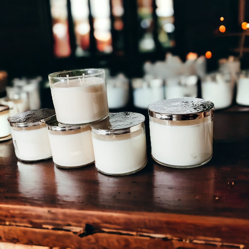 May include: A group of five white candles in glass jars with silver lids. The candles are arranged in a pyramid shape on a wooden surface.
