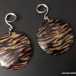 May include: A pair of round earrings with a dark brown and gold zebra stripe pattern. The earrings have a silver-colored, looped hook. The earrings are displayed on a dark surface.