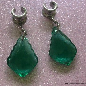 May include: A pair of silver ear cuffs with dangling green teardrop-shaped crystals. The crystals have a sparkly, glittery finish.