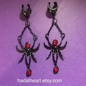 May include: A pair of black metal ear plugs featuring a dangling spider design. Each spider has a red teardrop-shaped gemstone on its back and a matching red teardrop charm hanging from the bottom. The text hadalheart.etsy.com is at the bottom.