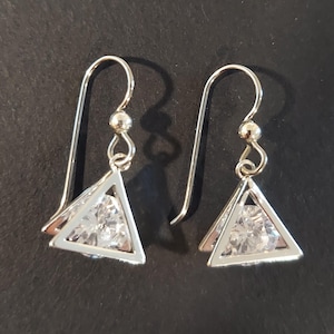 May include: A pair of silver-toned drop earrings featuring a geometric triangular frame. Each earring contains a clear, faceted, pyramid-shaped stone suspended within the center of the metal triangle, attached to a simple curved ear wire hook.