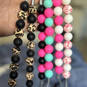May include: Five phone charms with different bead patterns. The charms are made of silicone beads in black, white, pink, and turquoise. The beads are strung on a silver chain with a silver hook at the end.