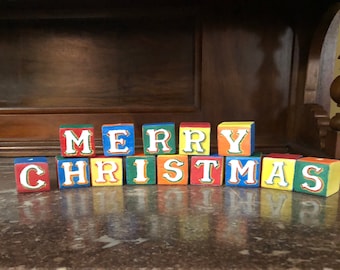 May include: Wooden blocks spelling out "Merry Christmas" in white letters. The blocks are painted in red, yellow, blue, and green.