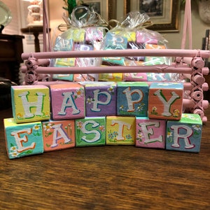 May include: Wooden blocks spelling out "Happy Easter" in pastel colors with white daisy accents. The blocks are arranged in a row on a wooden surface.