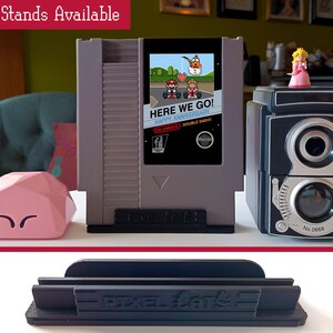 May include: Three different types of stands for video game cartridges. The stands are black and gray and have the text "Pixel Stand" printed on them. The stands are designed to hold video game cartridges upright.