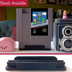 Puede incluir: Tres soportes de pl&aacute;stico negro para cartuchos de videojuegos. Los soportes est&aacute;n etiquetados con "Pixel-Art". Un soporte est&aacute; sosteniendo un cartucho de juego de Nintendo Entertainment System gris con una etiqueta que dice "Legendary Dad Happy Father's Day! A True Hero".