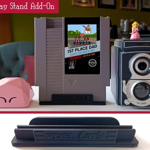 May include: A display stand add-on featuring a gray retro video game console design with a screen displaying a Father's Day message. A pink geometric object and a black and silver camera are also present. A black stand is at the bottom.
