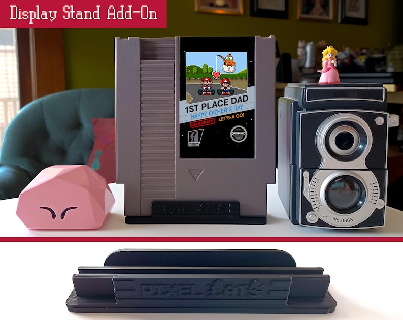 May include: A display stand add-on featuring a gray retro video game console design with a screen displaying a Father's Day message. A pink geometric object and a black and silver camera are also present. A black stand is at the bottom.