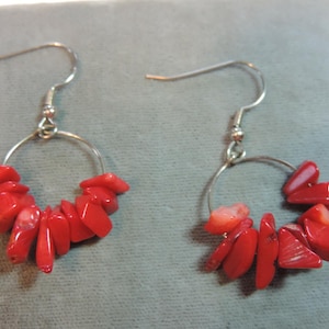 May include: A pair of dangle earrings featuring a silver-tone hoop design adorned with vibrant red coral chips. The earrings have a simple hook closure. The coral chips are irregularly shaped and add a pop of color.