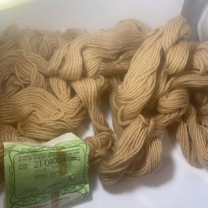 May include: A pile of tan-colored yarn skeins with a green and white label. The yarn is loosely arranged, showing the texture of the fibers. The label provides color and dye lot information.