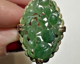 May include: Oval-shaped ring featuring a carved green jade design of flowers and leaves set in a gold-toned metal frame. The jade is a vibrant green color, and the ring has a detailed, ornate appearance. The ring is a piece of jewelry.