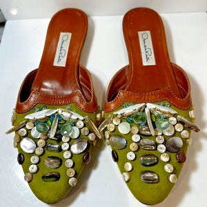 May include: A pair of olive green mule sandals with embellishments. The sandals feature a brown leather insole and trim, with decorative elements including iridescent shells, beads, and stones. The brand name "Oscar de la Renta" is visible on the insole.