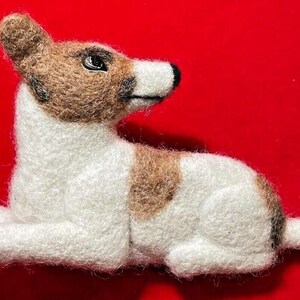 May include: A handmade felt dog figurine in a resting pose. The dog has a white body with brown patches on its back and head. The dog's head and ears are brown, with black eyes and nose. The background is a solid red.