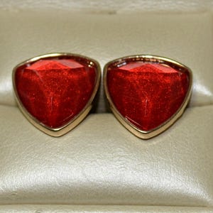 May include: A pair of red, triangular-shaped earrings with a gold-colored border. The earrings have a faceted, gem-like appearance and are set against a neutral, beige background.