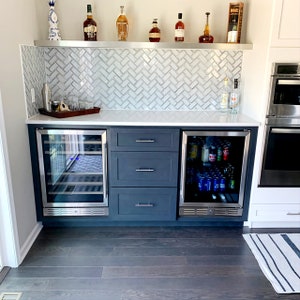 May include: A white countertop with two stainless steel mini-fridges, one with wine bottles and the other with canned beverages. The countertop is flanked by dark blue cabinets with silver drawer pulls. The countertop is also adorned with a stainless steel shelf with various bottles of liquor.