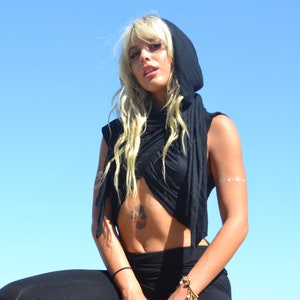 May include: A woman wearing a black hooded top and black trousers with ruffles at the bottom. She is sitting on a black stool in front of a swimming pool.