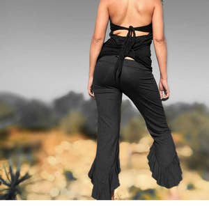 Könnte beinhalten: Eine Frau trägt ein schwarzes Halstuch-Top und eine schwarze Hose mit Rüschen am Saum. Die Hose ist locker sitzend und fließend. Die Frau steht auf einem Feld mit verschwommenem Hintergrund.