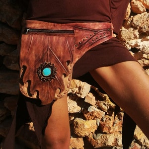 May include: A brown leather belt with a decorative leaf design and a turquoise stone accent. The belt is worn over a brown long-sleeved shirt.