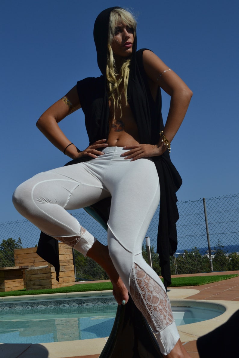 May include: A woman wearing a black hooded top with white pants and lace trim. She is standing by a pool with a fence in the background.