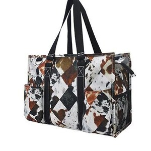 Op de afbeelding: Een grote tote bag met zwarte handvatten en een patchwork-ontwerp met bruine, witte en zwarte koeienhuidprint. De tas heeft meerdere vakken en een zijvak van gaas.