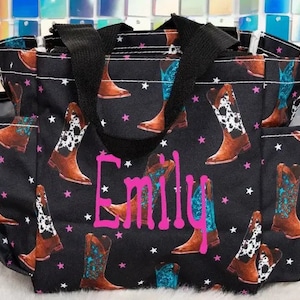Puede incluir: Bolsa de mano negra con un patrón de botas de vaquero y estrellas. La bolsa tiene dos asas negras y el nombre "Emily" está escrito en letras rosas en la parte delantera.