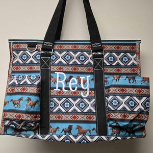 May include: A blue and brown tote bag with a geometric pattern and horse designs. The bag has a large pocket on the side and the name "Rey" is embroidered on the front.