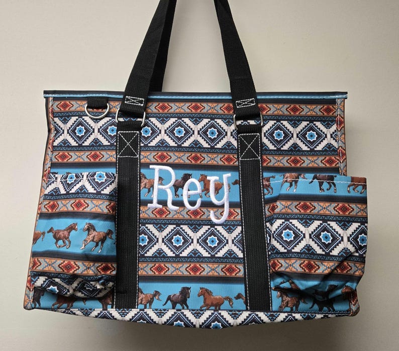 May include: A blue and brown tote bag with a geometric pattern and horse designs. The bag has a large pocket on the side and the name "Rey" is embroidered on the front.