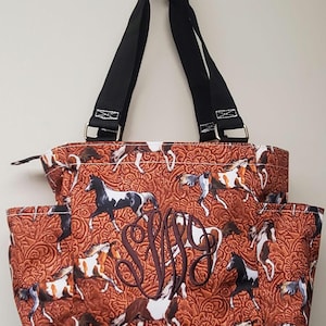 Könnte beinhalten: Eine braune und weiße Tote Bag mit Pferde-Print, mehreren Taschen und einem schwarzen Riemen. Die Tasche ist mit den Buchstaben "S.W." in braunem Garn bestickt.