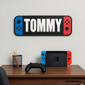 May include: A black wall sign with the name "TOMMY" in white, designed like a game console controller. Below, a game console, controller, and pencil holder sit on a wooden desk. The console has blue and red accents.