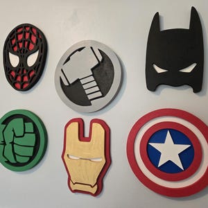 Könnte beinhalten: Eine Sammlung farbenfroher Holz-Superhelden-Embleme, darunter Spider-Man, Thors Hammer, Batman, Hulks Faust, Iron Mans Maske und Captain Americas Schild. Diese dekorativen Stücke sind an einer hellgrauen Wand befestigt.