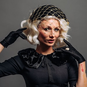Könnte beinhalten: Eine Frau in einem schwarzen Kleid mit Samtkragen und einem goldenen Netz-Hut mit Schleier. Sie trägt schwarze Handschuhe und hat ein Tattoo am Arm.