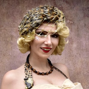 Könnte beinhalten: Eine Frau in einem Leopardenmusterkleid mit einem goldenen und schwarzen Hut und einer braunen Perlenkette. Das Kleid hat eine große Schleife vorne und eine goldene Brosche mit einem schwarzen Katzenmotiv. Der Text "Jezebel's Fascination" ist in der rechten unteren Ecke des Bildes zu sehen.