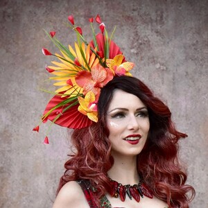 Puede incluir: Una mujer con cabello rojo lleva un tocado tropical vibrante con flores y follaje rojo, amarillo y naranja. También lleva un vestido floral rojo y verde y sostiene una taza tiki. La imagen tiene el texto "Jezebel's Fascination".