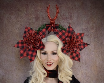 May include: A woman with blonde hair is wearing a red and black plaid headband with a red reindeer antler and poinsettia flower accents. The headband is decorated with green garland. She is also wearing a red and black plaid scarf.