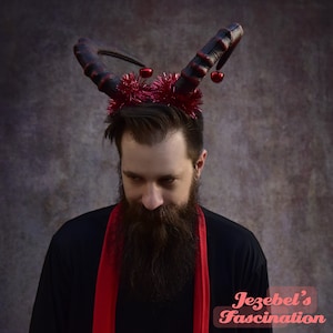 May include: A person wearing a black shirt and a red scarf with a red and black devil horn headband with red jingle bells. The text "Jezebel's Fascination" is visible in the lower right corner of the image.