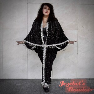May include: A woman wearing a black, star-patterned robe with silver fringe and a silver chain necklace. The robe has a long, flowing silhouette and is cinched at the waist. The woman is standing in front of a white tiled wall.