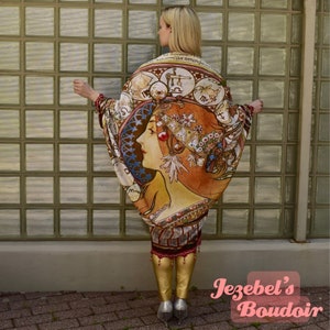 May include: A woman wearing a long, flowing, multi-colored kimono with a detailed illustration of a woman's face and zodiac symbols. The kimono is worn over gold leggings and the woman is standing in front of a glass block wall.