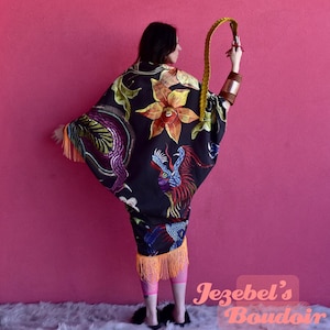 May include: A black kimono with colorful embroidered dragons, flowers, and fringe. The kimono is worn by a person with long dark hair. The background is a solid pink wall.
