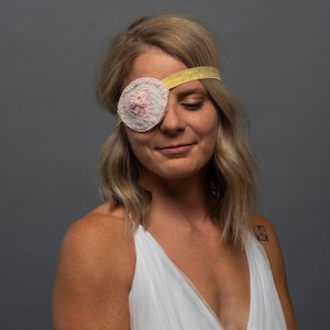 May include: A decorative eye patch with a heart design. The eye patch is light pink and white with a yellow glitter headband. The eye patch is worn on the left eye. The background is a solid gray.